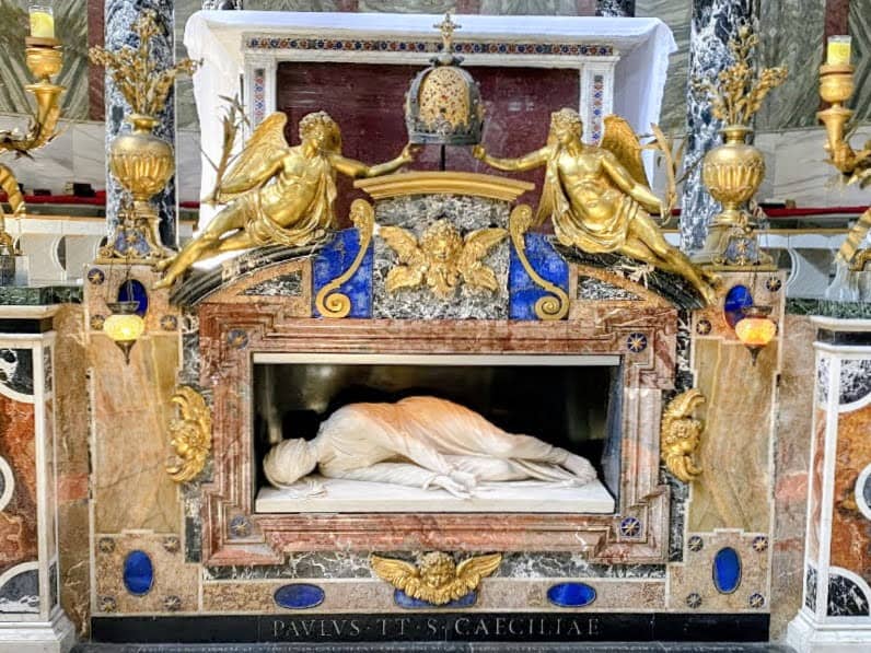  La famosa statua marmorea della santa, realizzata da Stefano Maderno nel 1599. Scultura sacra in marmo e oro, decorazioni barocche con angeli dorati e dettagli blu, all'interno di una chiesa storica, rappresenta un bassorilievo religioso con elementi artistici e spirituali.