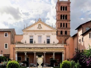 Antico edificio religioso con facciata in stile barocco, campanile in mattoni rossi, fontana al centro del giardino e piante verdeggianti, cielo nuvoloso, atmosfera storica e culturale.
