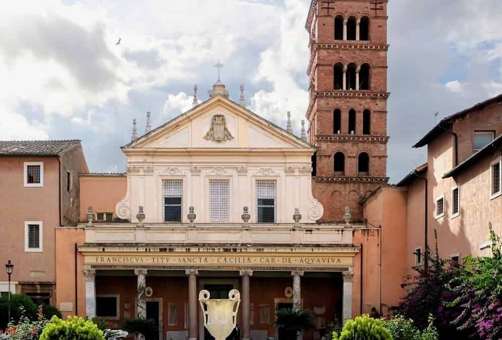 Antico edificio religioso con facciata in stile barocco, campanile in mattoni rossi, fontana al centro del giardino e piante verdeggianti, cielo nuvoloso, atmosfera storica e culturale.