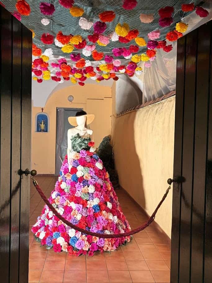 Colorata installazione floreale ispirata alle spose di lusso, decorata con un ampio vestito di fiori e un cappello di paglia, circondata da un soffitto di fiori colorati in un ambiente accogliente.