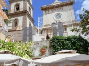 Campanile e chiesa storica in una pittoresca piazza con gazebo e persone al tavolo, cielo azzurro e nuvole bianche, atmosfera mediterranea, dettagli architettonici e vegetazione rigogliosa.