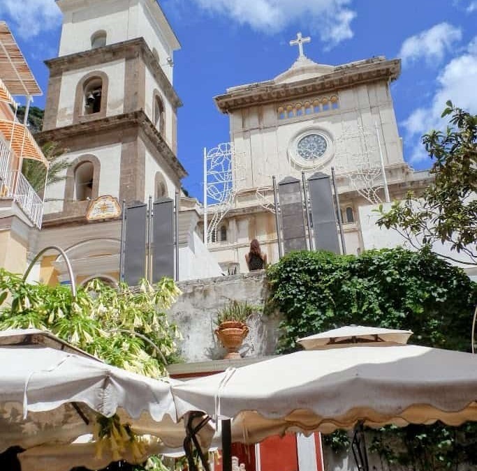 Campanile e chiesa storica in una pittoresca piazza con gazebo e persone al tavolo, cielo azzurro e nuvole bianche, atmosfera mediterranea, dettagli architettonici e vegetazione rigogliosa.