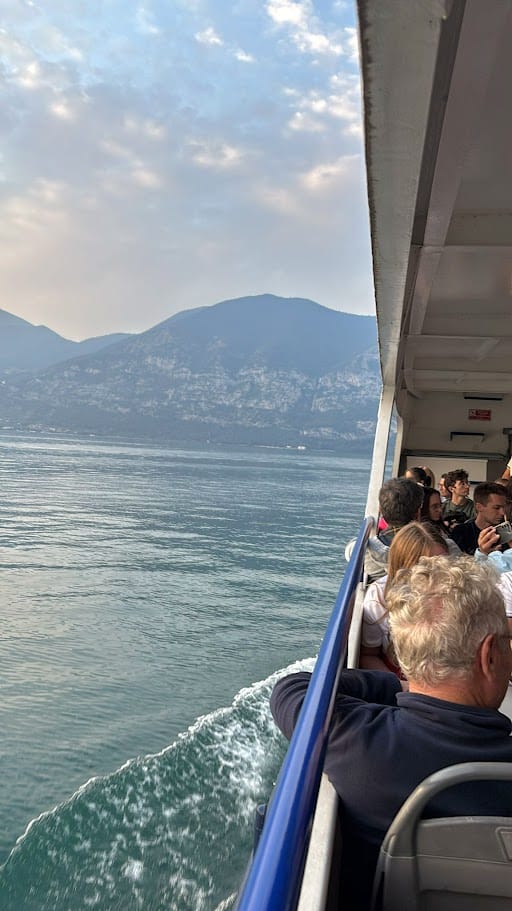 Passeggeri su un battello che naviga su un lago ai piedi di montagne, con cielo nuvoloso, atmosfera di viaggio e relax sulla barca.