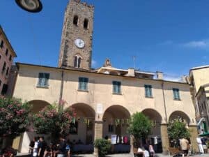 Splendida piazza storica nel centro di Town con le sue arcate e il campanile medievale, ambiente vivace con negozi, botteghe e passeggiata tra passeggini e persone.