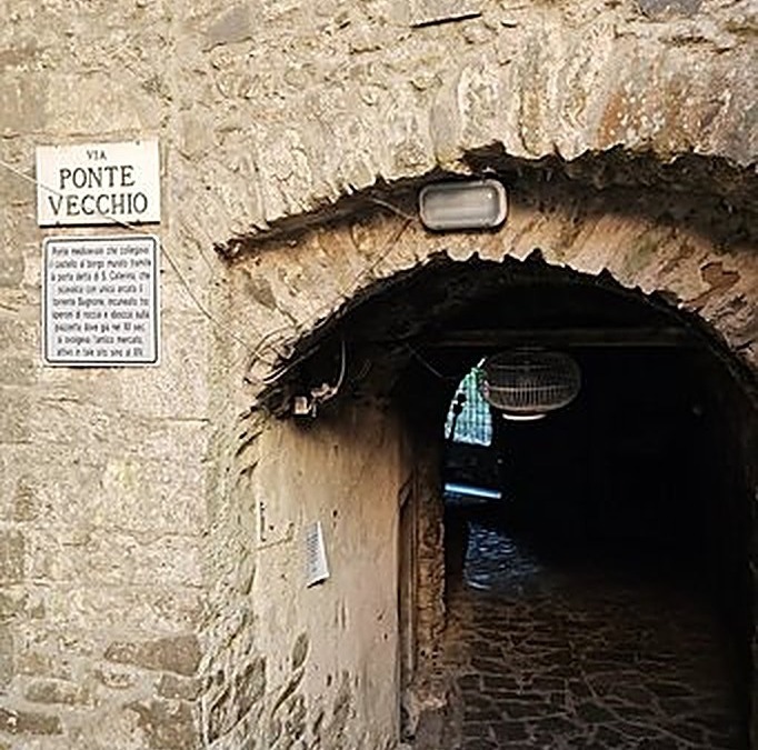 Ingresso di un passaggio storico in pietra con lampada, situato sotto un arco vicino a una strada antica, perfetto per esplorare il patrimonio culturale e architettonico italiano.