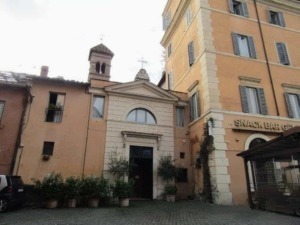 Vecchia chiesa nel centro di Roma, situata vicino a un edificio residenziale, con un piccolo cortile e piante, testimonianza di architettura storica italiana e ambientazione urbana.