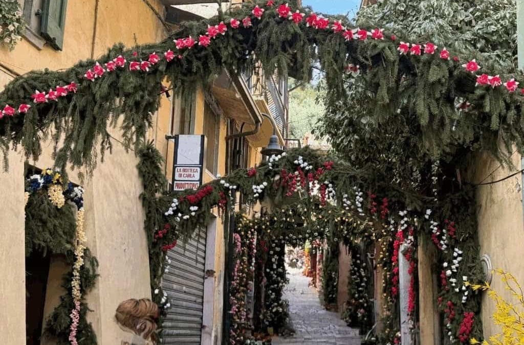 Fiori e decorazioni natalizie su un vicolo storico di una città italiana, con archi di piante verdi e fiori colorati, creando un'atmosfera festosa e accogliente nel centro urbano.