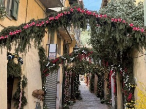 Fiori e decorazioni natalizie su un vicolo storico di una città italiana, con archi di piante verdi e fiori colorati, creando un'atmosfera festosa e accogliente nel centro urbano.
