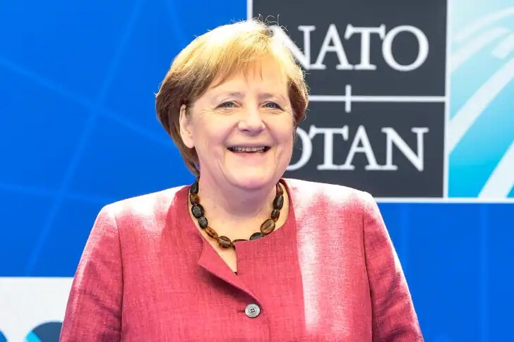 Angela Merkel in un evento internazionale, sorridente davanti a un backdrop NATO, rappresenta leadership politica e impegno diplomatico.