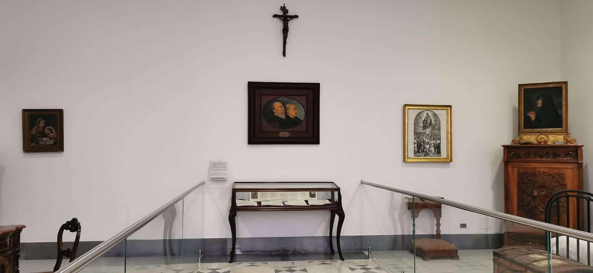 7 nuovi santi papa leone XIV da 4 continenti 3 San Bartolo Longo: Dalla Tenebra alla Luce. Ritratti storici e opere d'arte in una galleria museale di valore culturale. La parete presenta dipinti di personaggi religiosi e storici, con un crocifisso sopra, elementi tipici di un'esposizione artistica e religiosa.