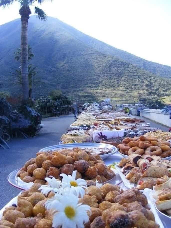 FOTO: Banchi di street food con vari snack tradizionali messicani, con lo sfondo di un paesaggio montano e arboreo, in una scena di mercato all'aperto con montagne, palma e cielo blu.