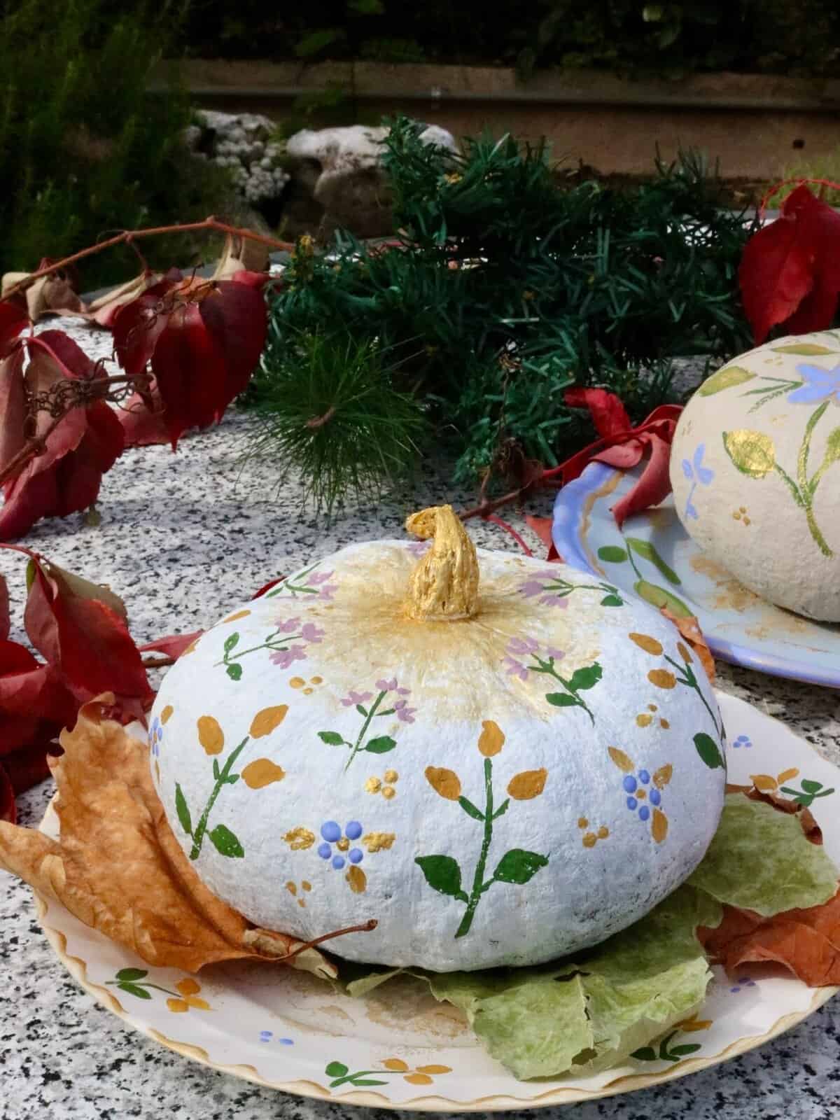 Orsara Festa della zucca, la festa della luce 3 Zucca decorata con motivi floreali in giallo, verde e blu, posta su un piatto con foglie secche, decorata con vernice dorata sul gambo, ambiente autunnale con foglie e piante verdi sullo sfondo.