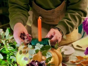 Ampio di una persona che crea una composizione artistica con zucche, foglie e fiori, utilizzando elementi naturali e decorativi per decorare uno spazio, con attenzione ai dettagli e alla creatività.