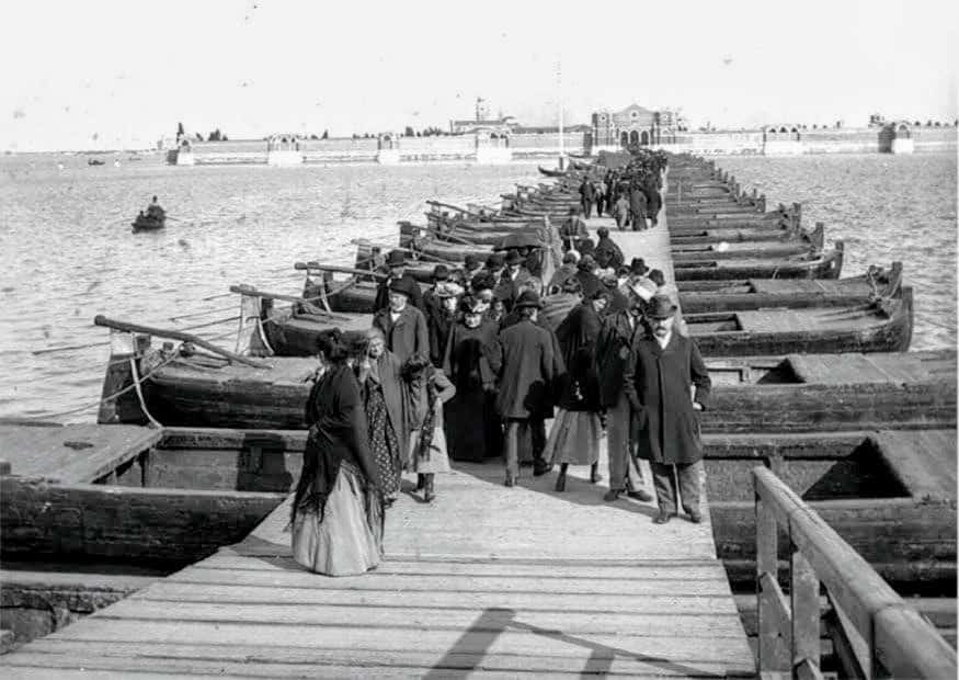 Immagine storica del ponte votivo fatto di barche con cittadini in abiti d'epoca, rappresenta la vita quotidiana nel passato marittimo.