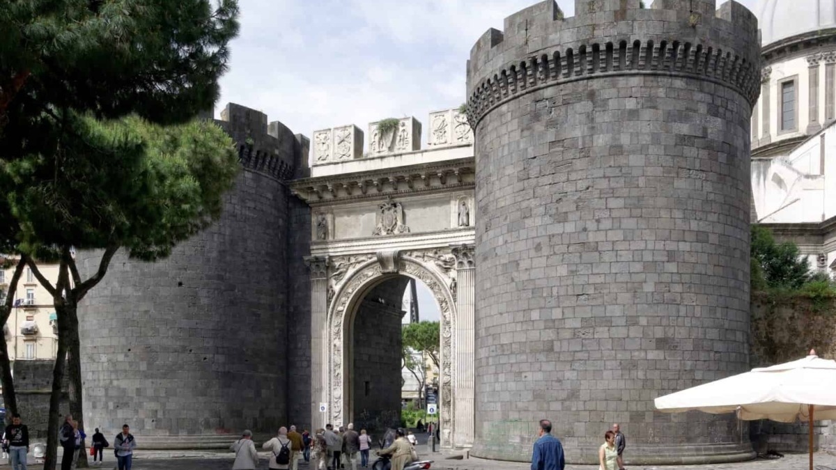 Antica porta medievale di Napoli con torri di pietra, nel cuore della città, popolata da passanti e motoveicoli.