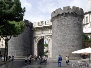 Antica porta medievale di Napoli con torri di pietra, nel cuore della città, popolata da passanti e motoveicoli.