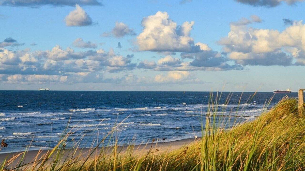 Prenota ora le tue vacanze: Tramonto sulla spiaggia con onde e cielo nuvoloso, vista panoramica del mare e delle dune di sabbia con erba alta, atmosfera tranquilla e paesaggio naturale.