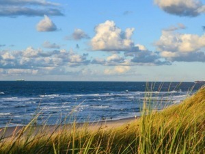 Prenota ora le tue vacanze: Tramonto sulla spiaggia con onde e cielo nuvoloso, vista panoramica del mare e delle dune di sabbia con erba alta, atmosfera tranquilla e paesaggio naturale.