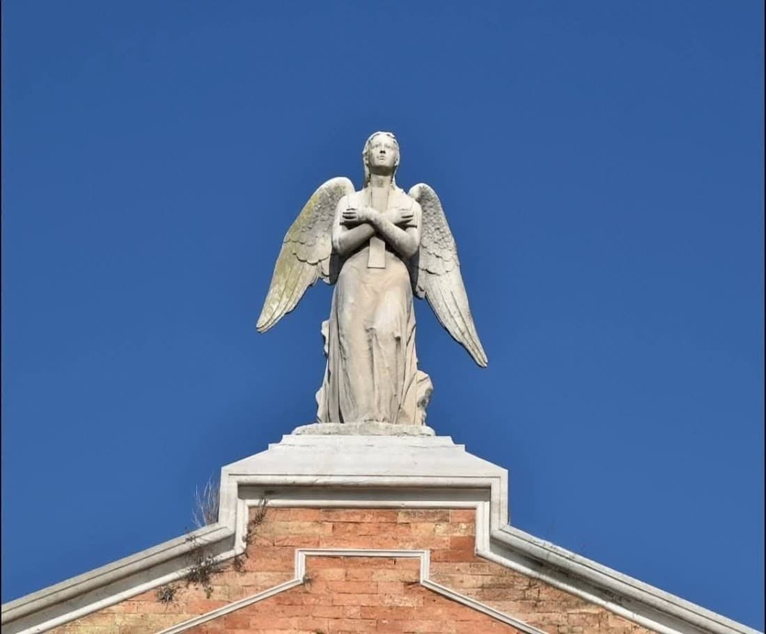 Statua di un angelo con ali in marmo bianco posizionata sopra un edificio in mattoni rossi, cielo azzurro sullo sfondo, vista dall'alto, simbolo di speranza e protezione, elementi architettonici classici, decorazione religiosa.
