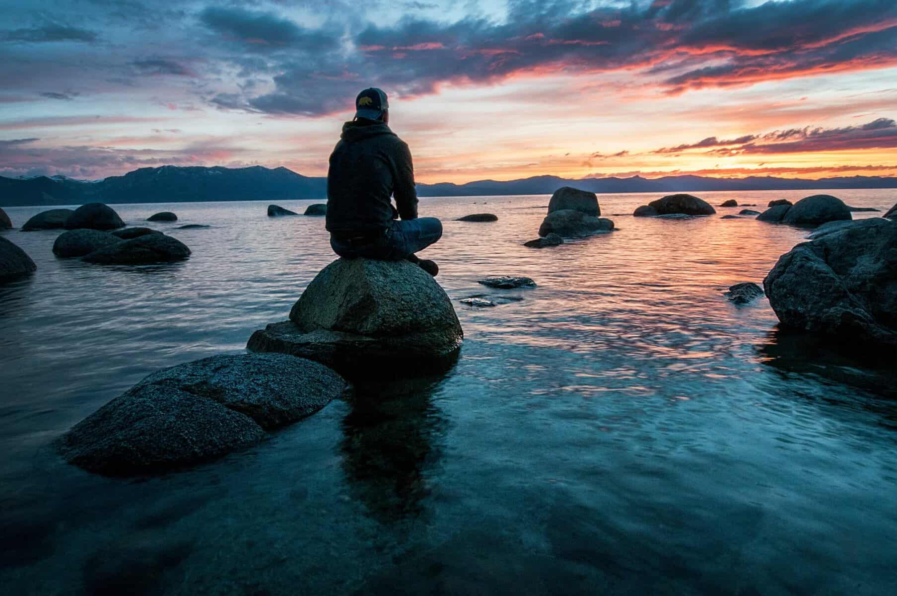 Riflesso di un tramonto sul lago con persona seduta su una roccia, paesaggio naturale, silenzioso e sereno, ideale per articoli su natura, relax e mindfulness.