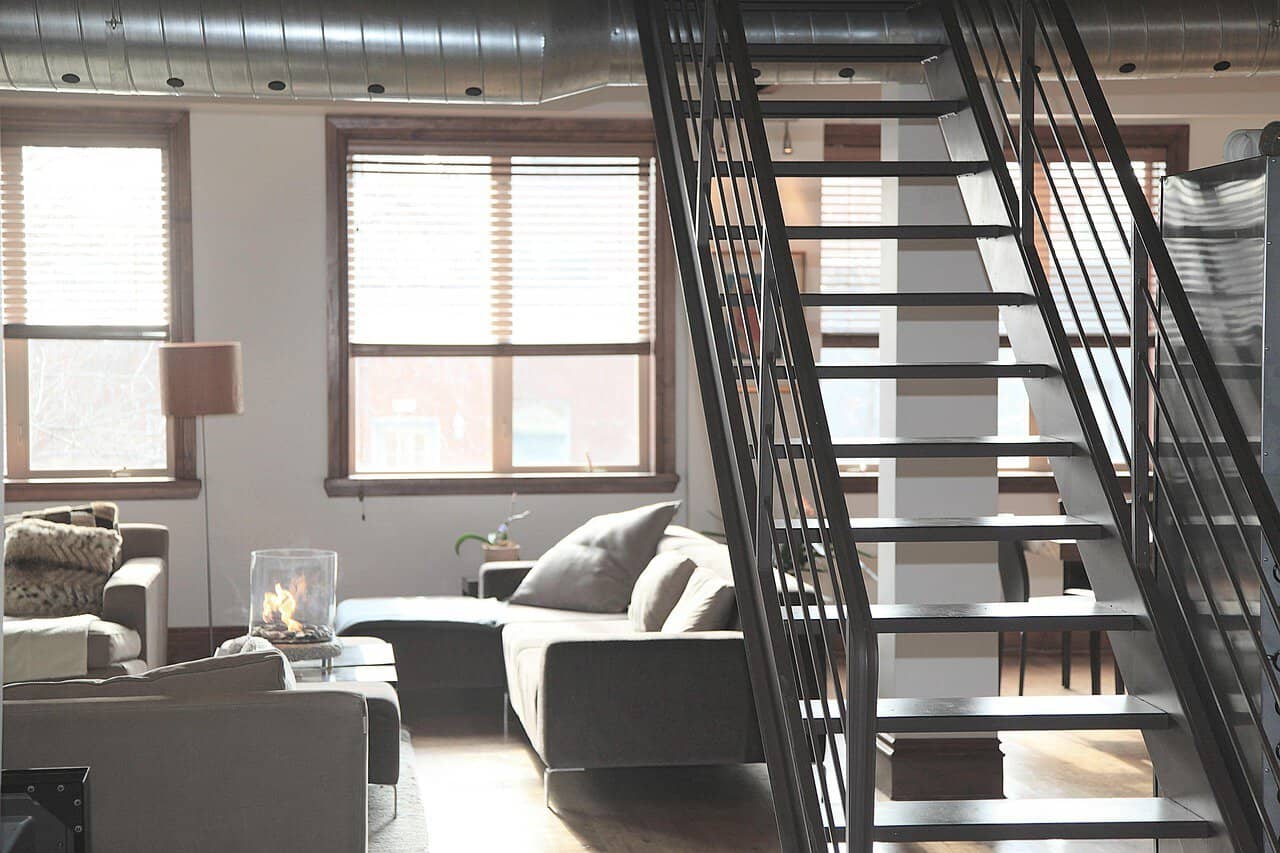 Moderno salotto con grandi finestre e pareti bianche, arredato con divani, lampada da terra e una scala in metallo che collega i piani superiori, in stile industrial chic.