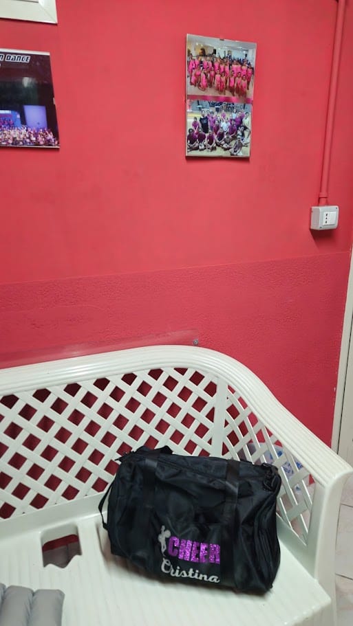 Borsa nera con nome "GFEAR Cristina" su panca di attesa in studio di fisioterapia. Pareti rosse con foto di gruppo di atleti di ginnastica, ambiente motivante e accogliente.