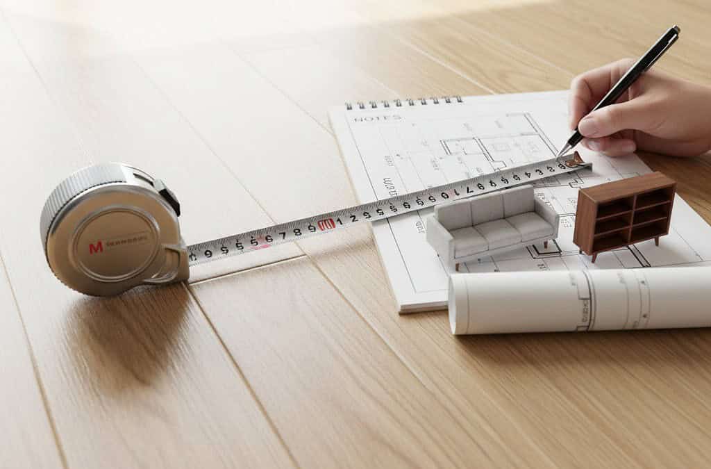 Alt text: Persona che disegna un piano di arredamento su blueprint con mini mobili, illuminato dalla luce naturale di una finestra. Utensili di progettazione come metro, carta e matita sono visibili.