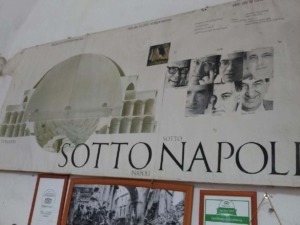 Immagine di un grande cartellone pubblicitario con il testo "SOTTO NAPOLI" e immagini di volti umani e architetture napoletane.