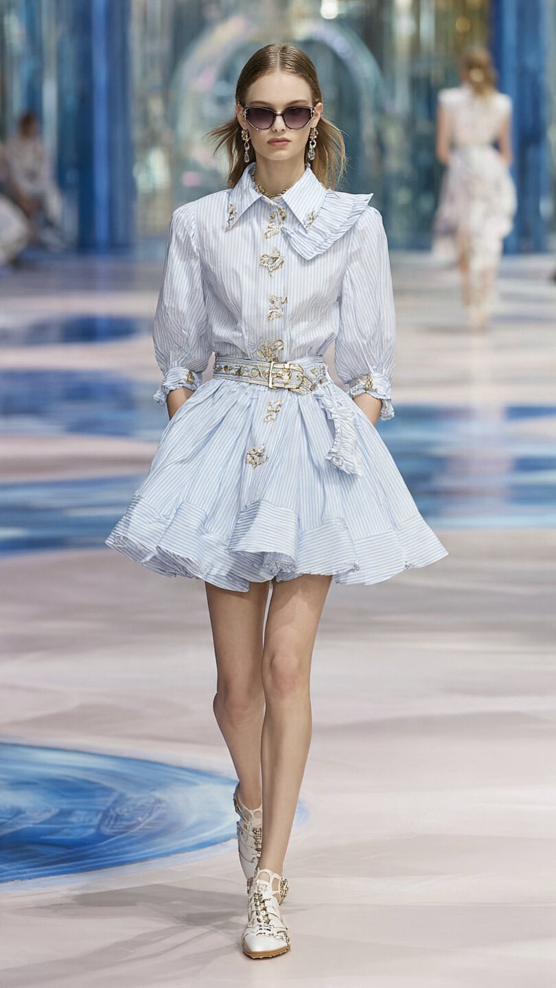 Quali sono i colori moda donna inverno 3 Modella indossa un abito estivo bianco con dettagli in pizzo e cintura, sfilando in passerella durante una fashion week.