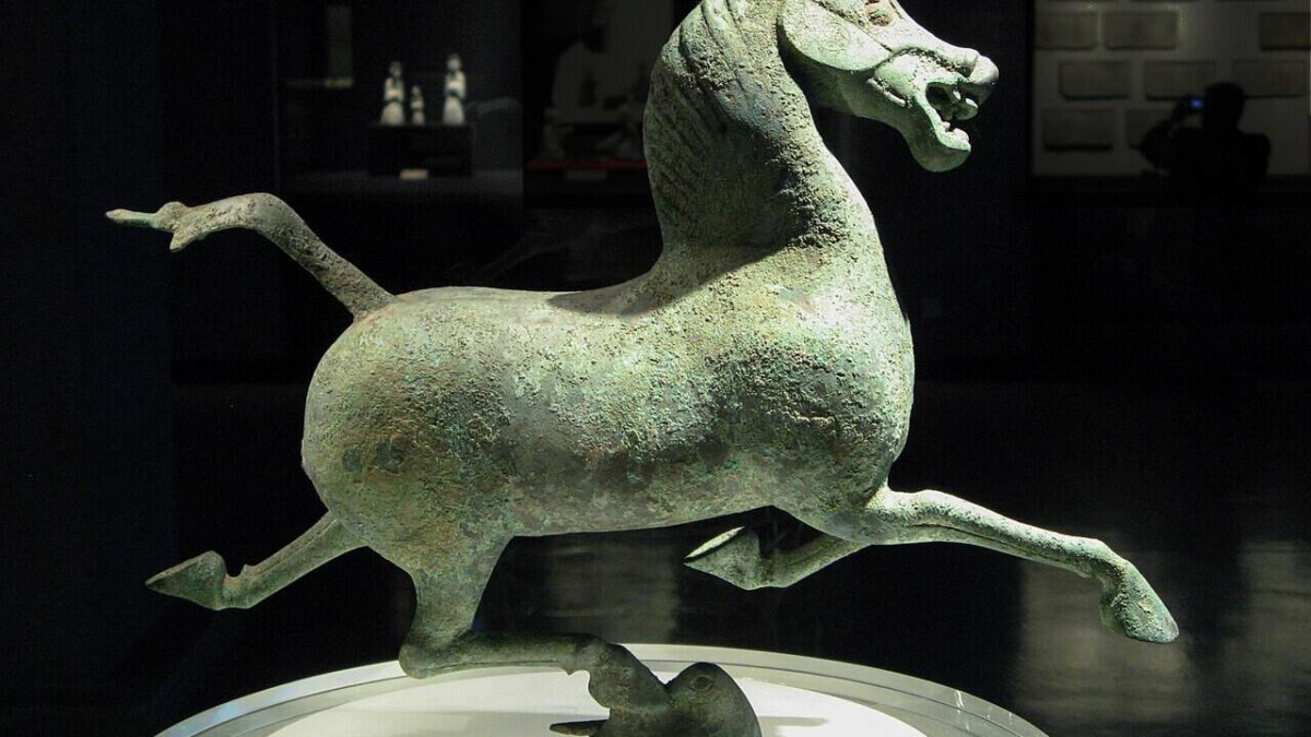 Statua di un cavallo in bronzo antico, esposta in un museo, con dettagli evidenti sulla superficie e postura dinamica, simbolo di arte e storia antica.