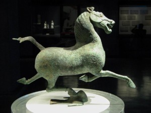 Statua di un cavallo in bronzo antico, esposta in un museo, con dettagli evidenti sulla superficie e postura dinamica, simbolo di arte e storia antica.