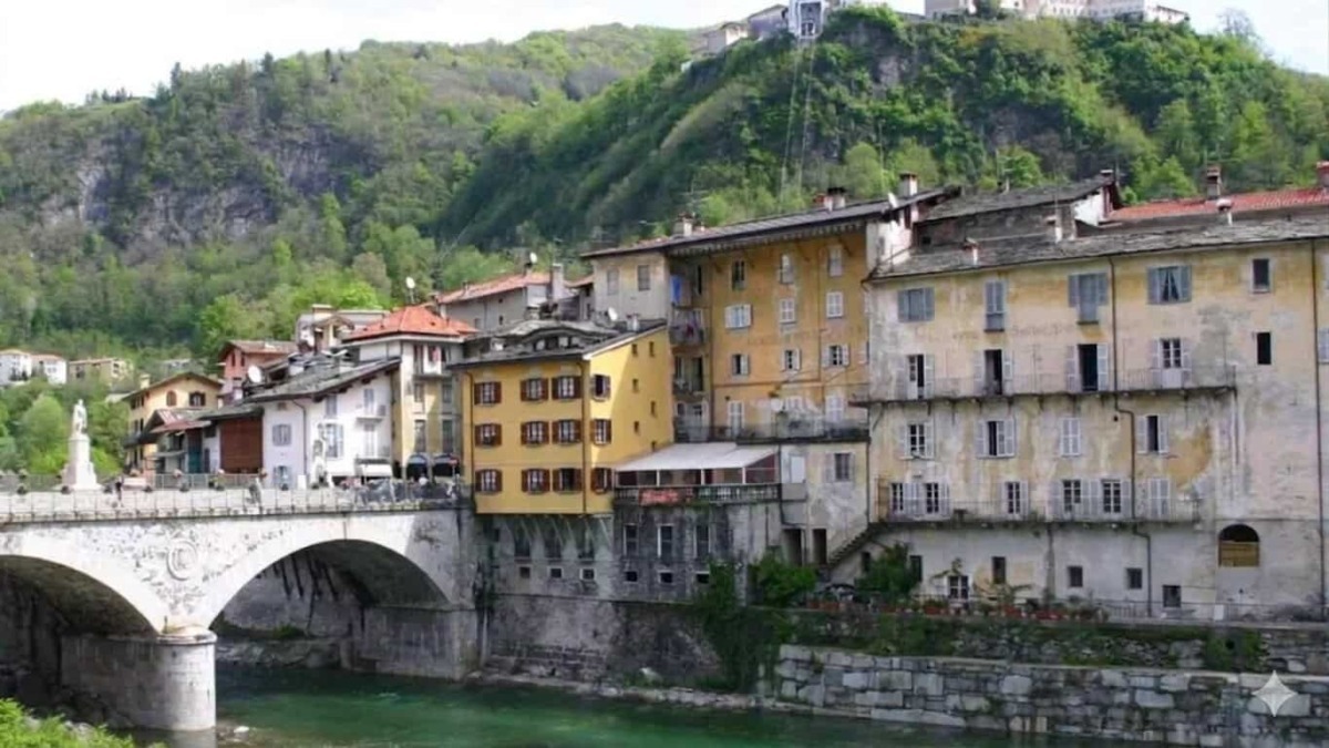 Vista di un quartiere antico con edifici colorati e un ponte in pietra che attraversa un fiume, circondato da colline verdi e natura rigogliosa, rappresentando un affascinante esempio di architettura.