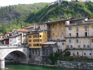Vista di un quartiere antico con edifici colorati e un ponte in pietra che attraversa un fiume, circondato da colline verdi e natura rigogliosa, rappresentando un affascinante esempio di architettura.