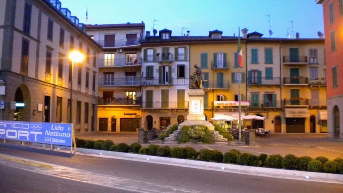Vista di una piazza italiana con monumento al centro, edifici storici con balconi e negozi, illuminata da luci serali, atmosfera tranquilla e caratteristica del centro città.