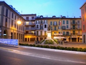 Vista di una piazza italiana con monumento al centro, edifici storici con balconi e negozi, illuminata da luci serali, atmosfera tranquilla e caratteristica del centro città.