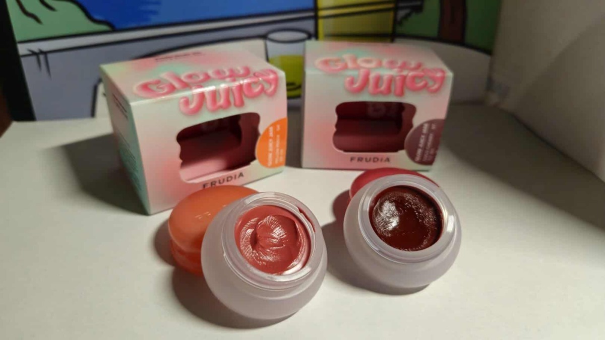 Due vasetti di crema labbra colorata, uno in tonalità rosa e l'altro in rosso scuro, posizionati su una superficie bianca con sfondo di scatole di prodotti di bellezza.