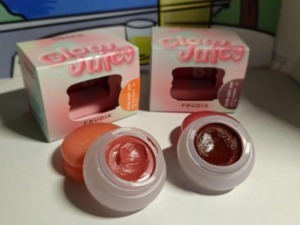Due vasetti di crema labbra colorata, uno in tonalità rosa e l'altro in rosso scuro, posizionati su una superficie bianca con sfondo di scatole di prodotti di bellezza.
