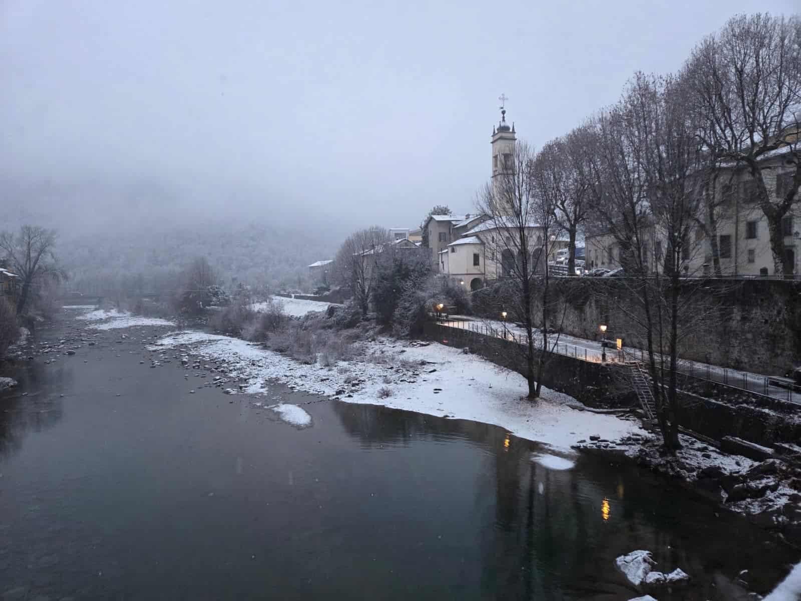 Paesaggio invernale con il fiume e il centro storico di Varallo Sesia, caratterizzato da edifici storici, una chiesa con torre e alberi ricoperti di neve, ideale per scoprire le attrazioni della zona.
