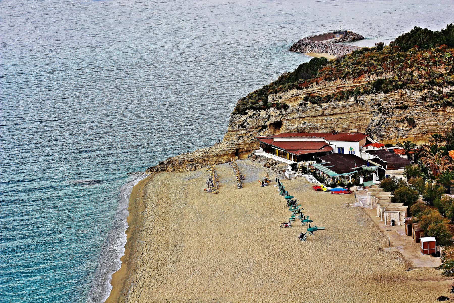 Spiaggia con sabbia dorata e scogliere, vista sul mare cristallino tra Puglia e Calabria, con stabilimenti balneari e vegetazione mediterranea.