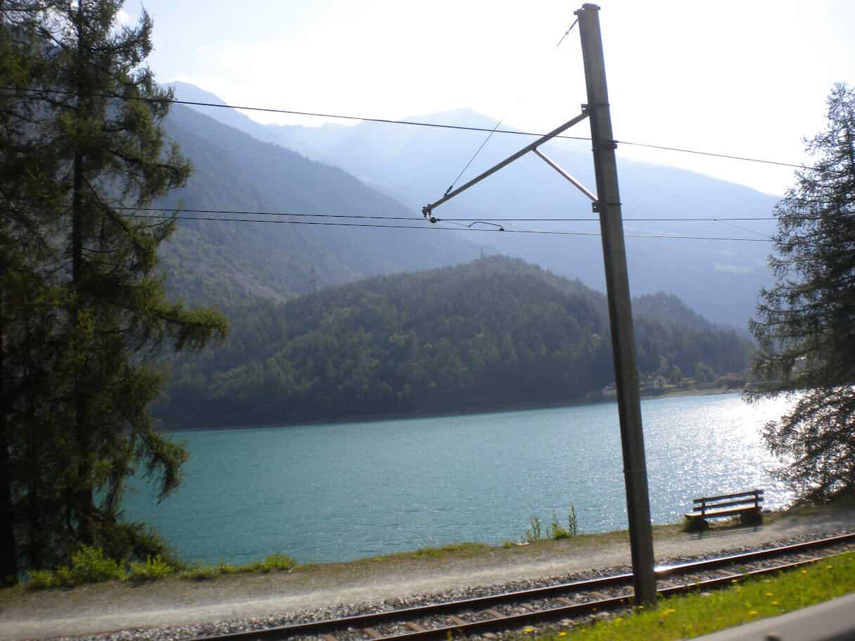 Treno che attraversa un paesaggio naturale con lago e montagne, ideale per appassionati di viaggi e natura. Vista tranquilla e suggestiva di un ambiente alpino con acqua cristallina e vegetazione rigo.