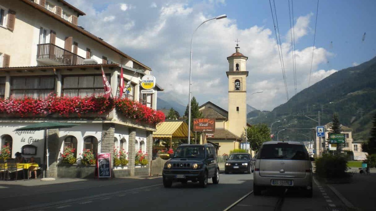 Vista del centro di Ortisei con chiesa e montagne sullo sfondo, strada trafficata e negozi fioriti, in una giornata soleggiata.
