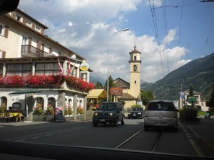 Vista del centro di Ortisei con chiesa e montagne sullo sfondo, strada trafficata e negozi fioriti, in una giornata soleggiata.