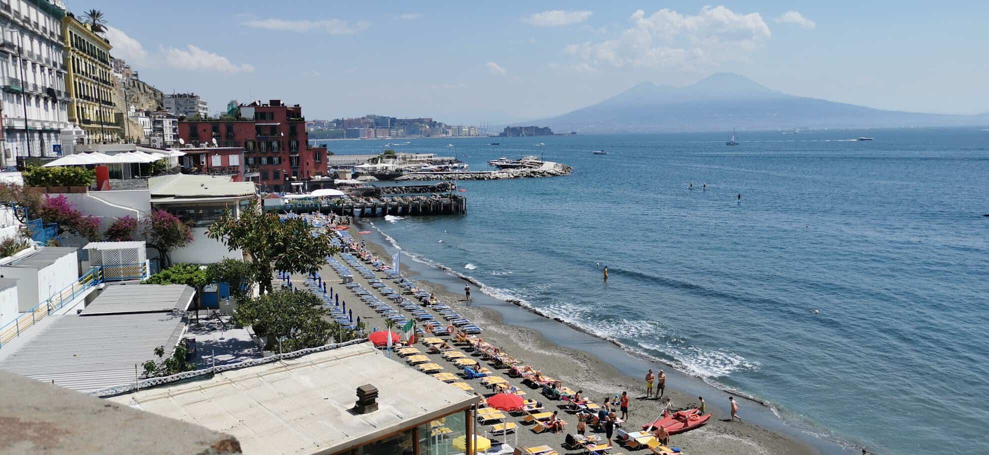 Spiaggia di Napoli con ombrelloni colorati, mare calmo e vista sul Vesuvio sullo sfondo, atmosfera estiva e turistica.