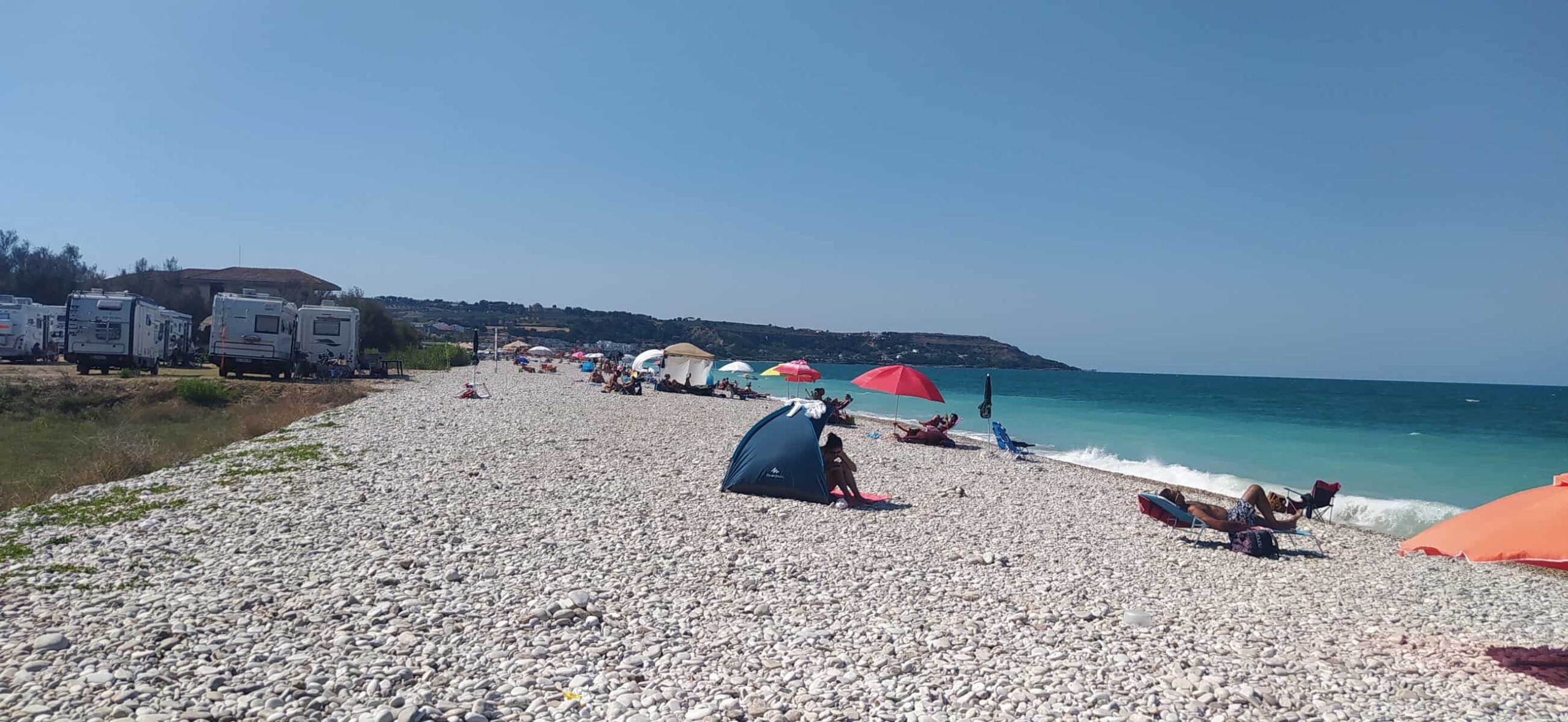 Spiaggia di ciottoli con ombrelloni colorati, lettini e persone che si rilassano vicino al mare in una giornata soleggiata.