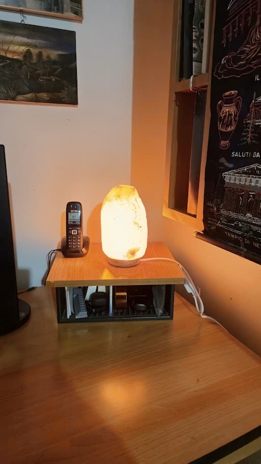 A cosa serve la lampada di sale, usi e benefici per purificare l'aria in casa 2 Lampada di sale himalaya: Lampada di sale accesa su tavolo in ambiente domestico, con telefono e decorazioni sullo sfondo, ideale per purificare l'aria in casa.