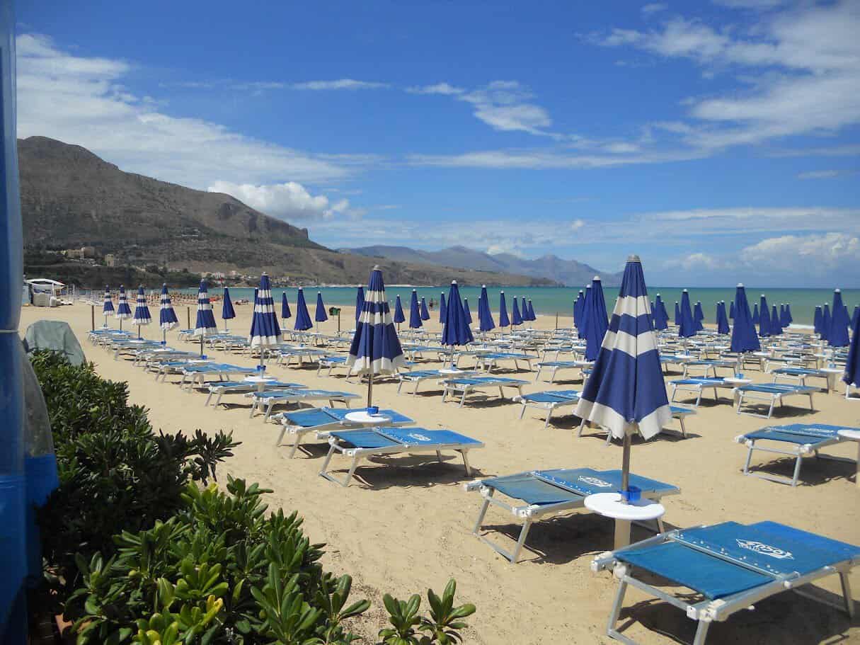 Sicilia. Spiaggia con numerosi ombrelloni blu e lettini sulla sabbia, vista sul mare e montagne sullo sfondo, cielo sereno con alcune nuvole.