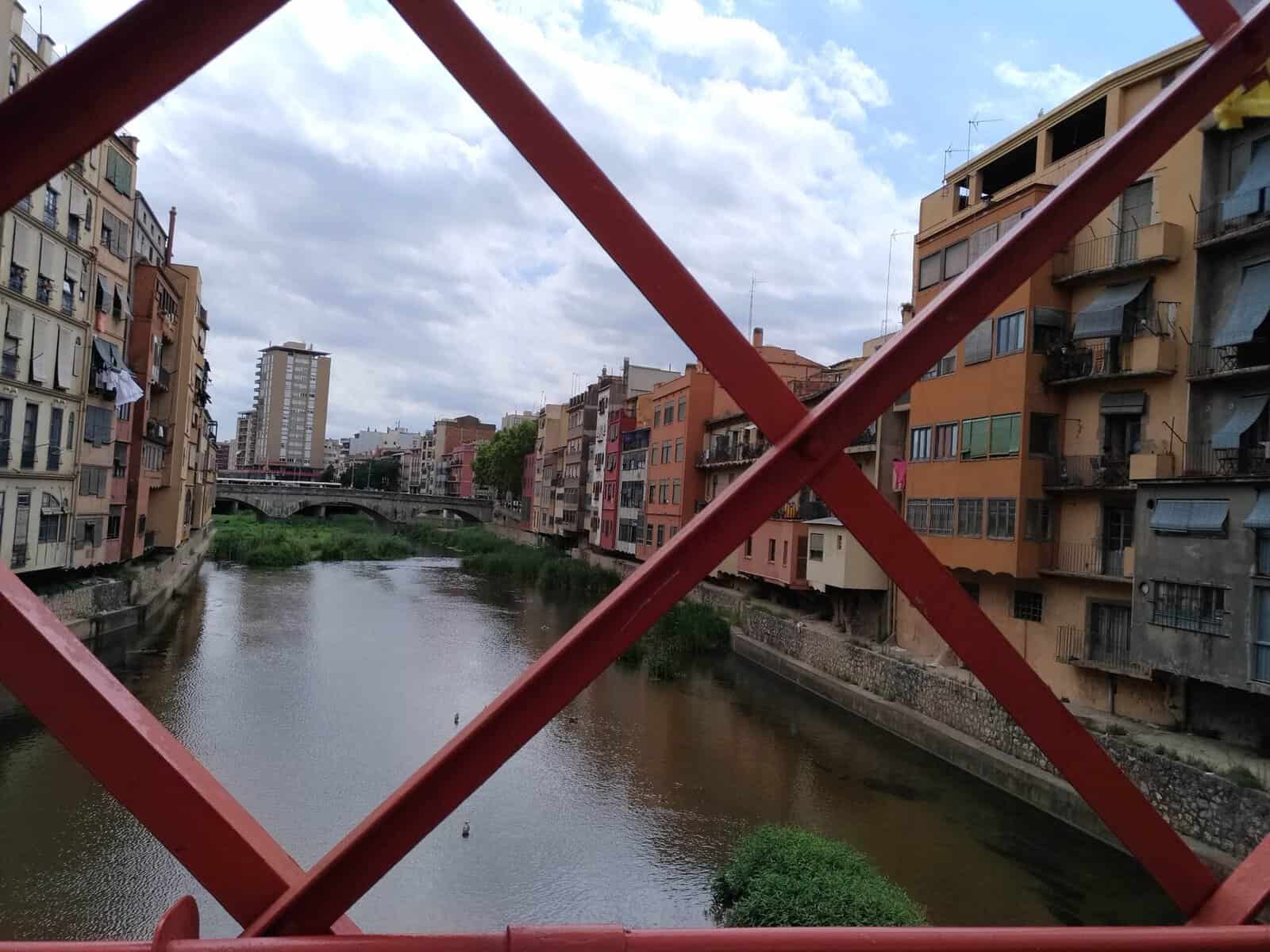 Cosa vedere a Girona in un giorno 5 Immagine di un fiume cittadino con edifici residenziali e un ponte rosso in primo piano, cielo nuvoloso, in un contesto urbano.
