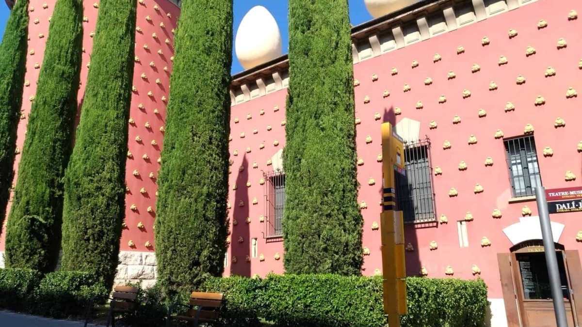 Edificio rosa con decorazioni gialle e grandi sfere bianche sul tetto, circondato da alberi di alto fusto e panchine lungo il marciapiede.