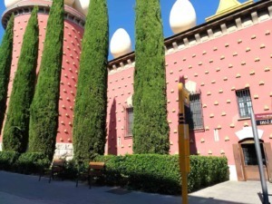 Edificio rosa con decorazioni gialle e grandi sfere bianche sul tetto, circondato da alberi di alto fusto e panchine lungo il marciapiede.