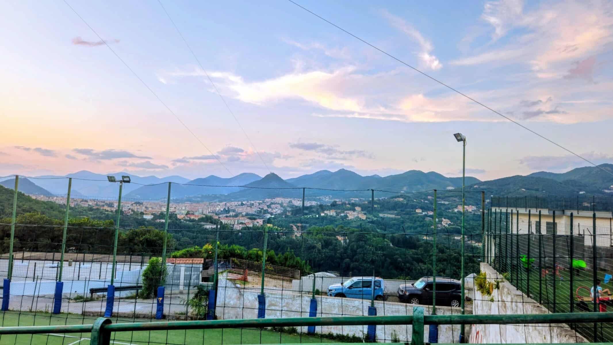 Vista panoramica di una città circondata da montagne con cielo al tramonto, lampioni e parcheggio in primo piano.